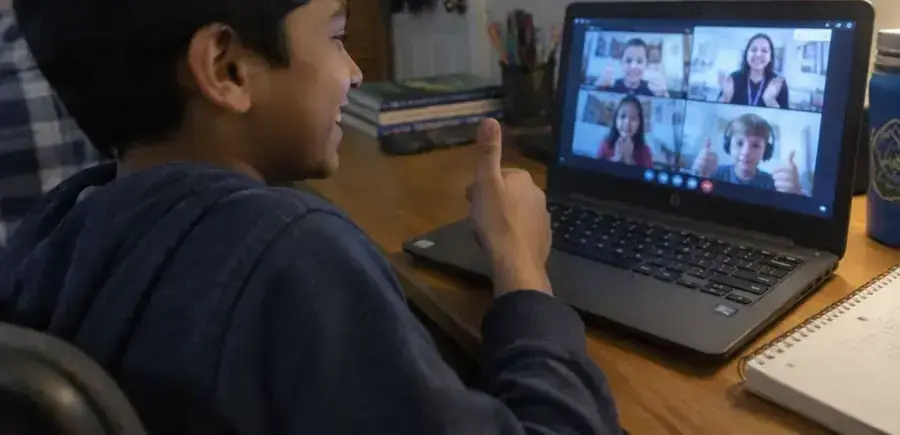 A boy waves hello to his live online class while his mother smiles beside him