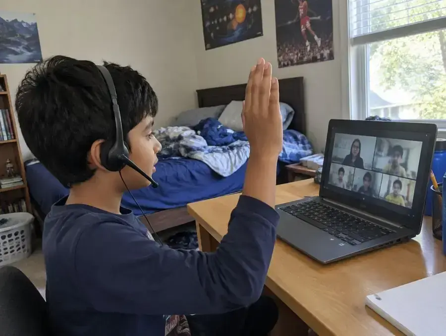 A boy in a headset raises his hand in a live online class at his desk