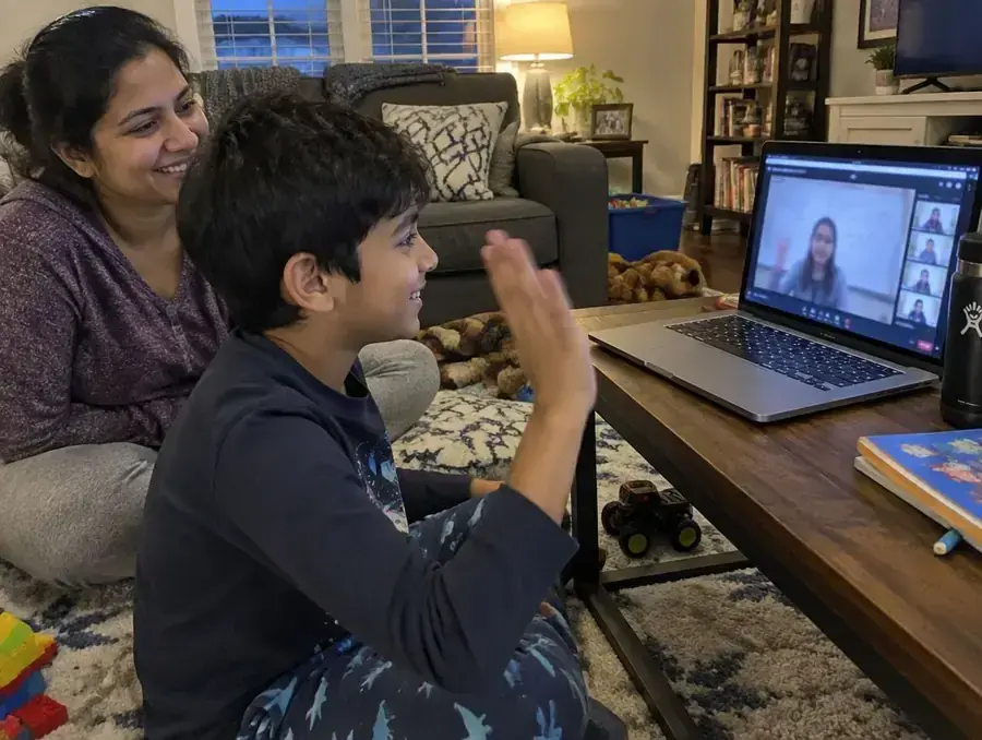 A boy waves hello to his live online class while his mother smiles beside him