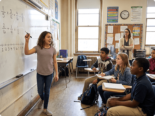The same girl at the whiteboard of her school classroom, marker in hand, explaining her working to her classmates while the teacher stands back.