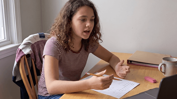The same girl at the same desk, sitting upright with her pencil raised, explaining her math working aloud.