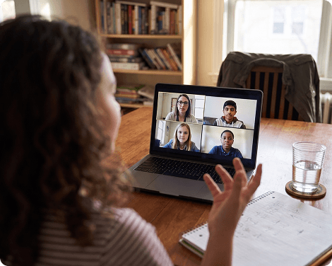Over her shoulder: the laptop shows her teacher and three classmates of the same age, all listening, while she gestures mid-explanation.