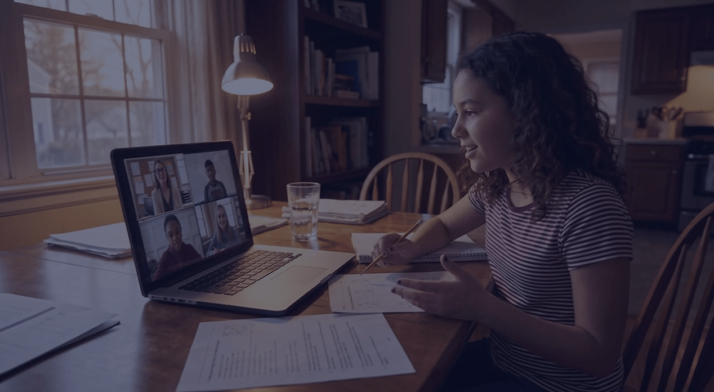 A girl of about twelve at the kitchen table explaining her working to her live math class, her teacher and three classmates on the laptop screen.